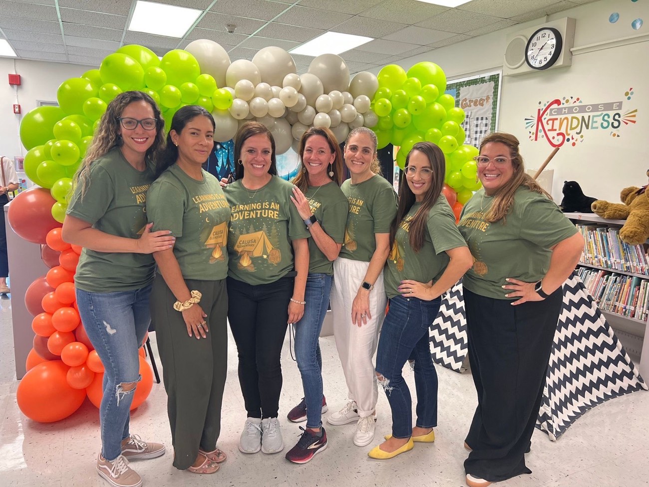 The seven Calusa PTA board members smiling together in matching 'Learning is an Adventure' shirts at the school media center