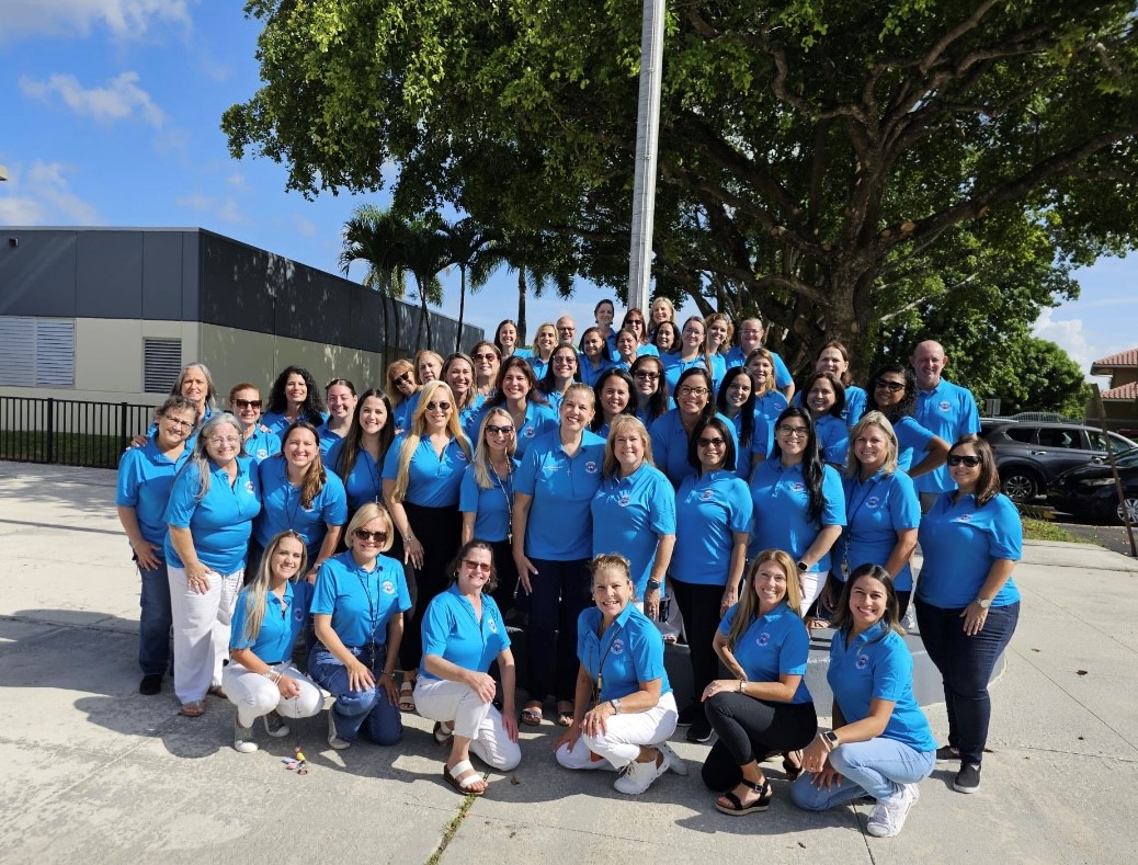 The full Calusa Elementary faculty and staff in matching blue polos, gathered under the big tree in front of the school